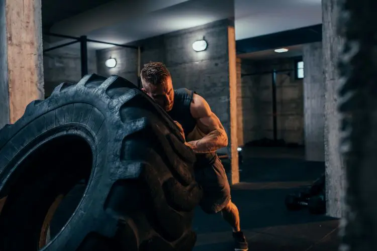 Muscular man in a dimly lit basement-like room pushes himself against a giant tire as he lifts it from the floor.